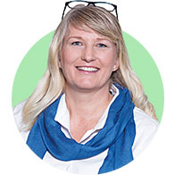 A woman with long blonde hair, wearing glasses on her head, a white shirt, and a blue scarf, set against a green circular background. Her face is intentionally blurred.