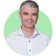 A man with gray-slicked hair wearing a white button-up shirt stands against a green circular background. His face is blurred, and no visible text or branding is present.