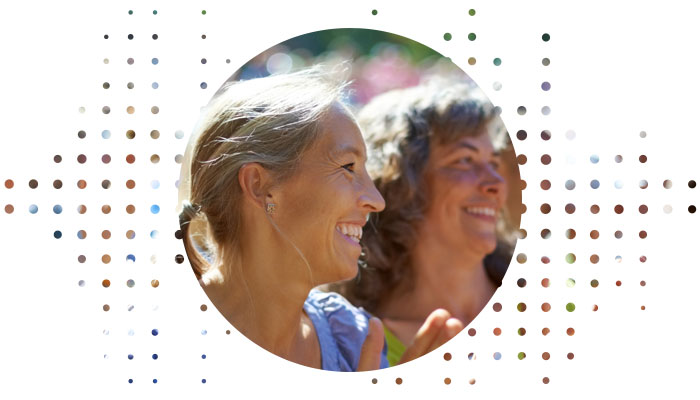 A woman wearing a discreet hearing aid behind her ear listens attentively outdoors on a sunny day, surrounded by blurred individuals. The dotted design frames the image dynamically.