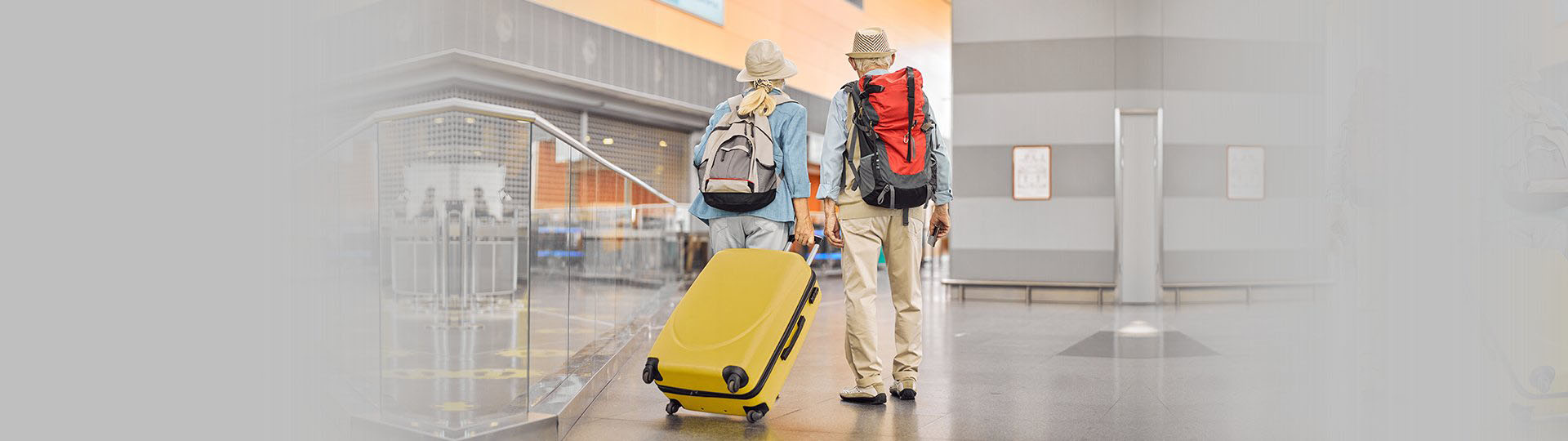 Older couple traveling through an airport terminal with backpacks and a bright yellow suitcase.