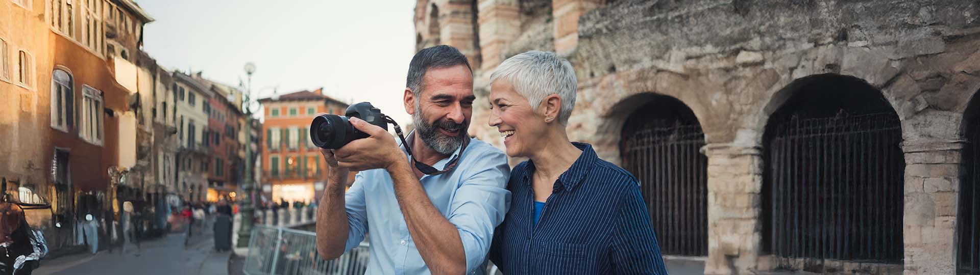 Couple smiling and leaning toward each other while holding a camera outdoors near historic architecture.