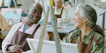 man and woman smiling at each other during an art class, with paintings and supplies in the background