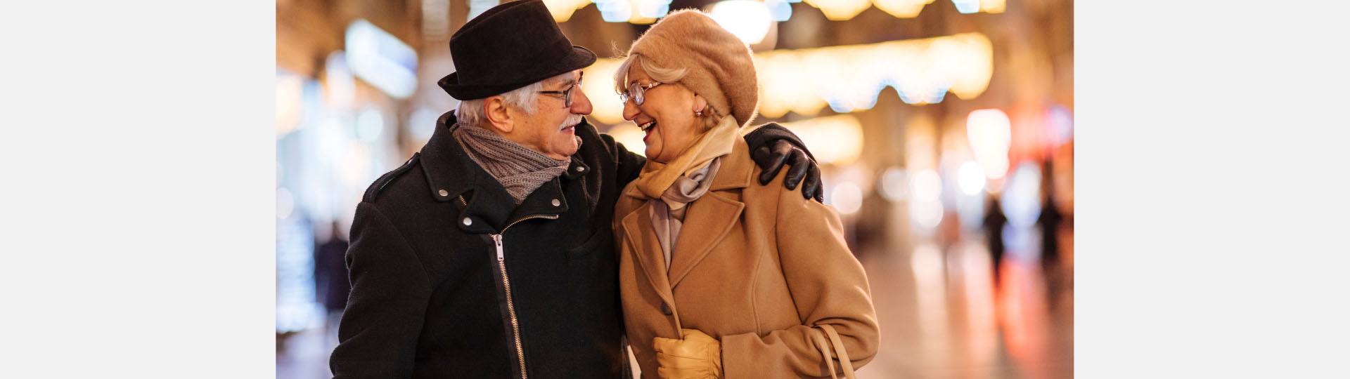 A couple in winter coats smiling and embracing outdoors at night with blurred festive lights illuminating the background.