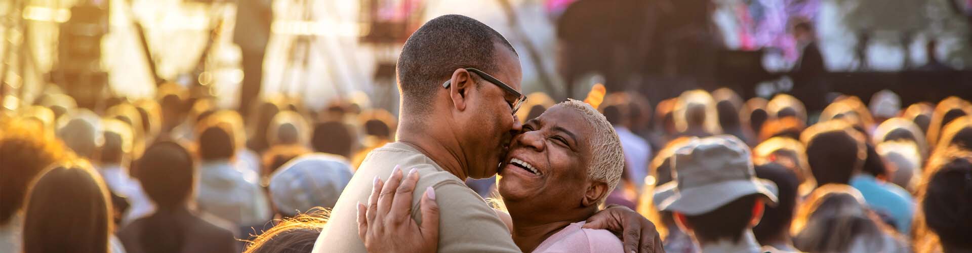 A couple sharing a joyful embrace surrounded by a crowd at an outdoor concert, with golden sunlight enhancing the lively atmosphere.