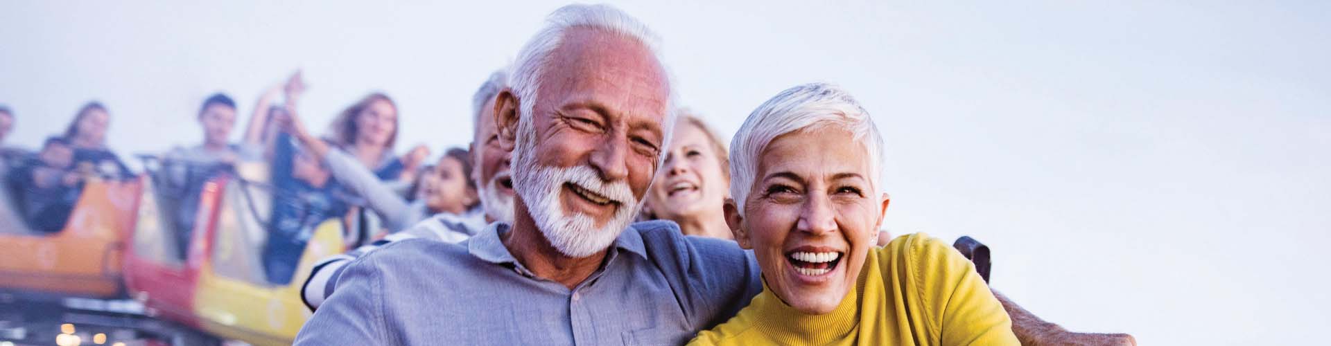Older couple laughing and enjoying a roller coaster ride with a crowd in the background.