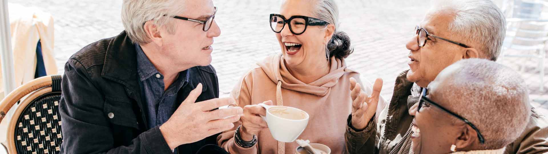 Older adults smiling and talking while sharing coffee at an outdoor café.