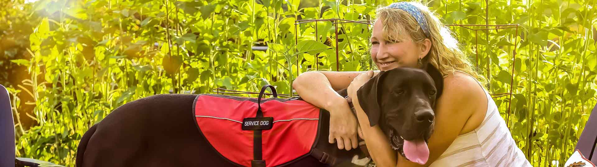 hearing assistance service dog in a red vest outdoors, leaning against a smiling person in a garden setting