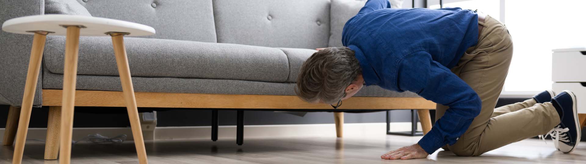 man looking for a hearing aid on the floor underneath a sofa