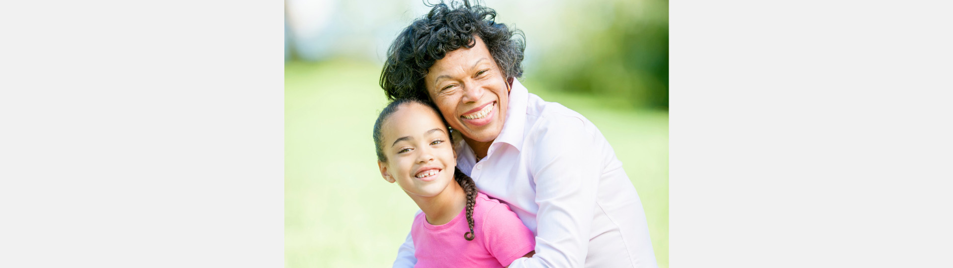 Woman smiling with a child outdoors on a sunny day, surrounded by lush greenery.