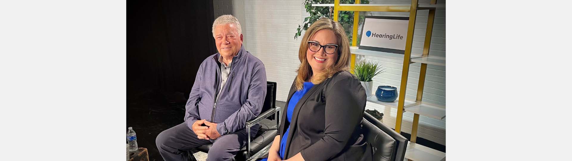 William Shatner and Dr. Rose seated in a modern studio with a HearingLife logo displayed among shelves and plants in the background.