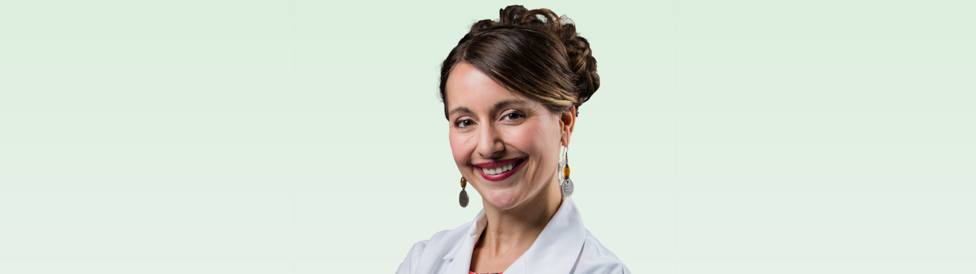 Smiling woman with styled hair and earrings, standing against a light green background.