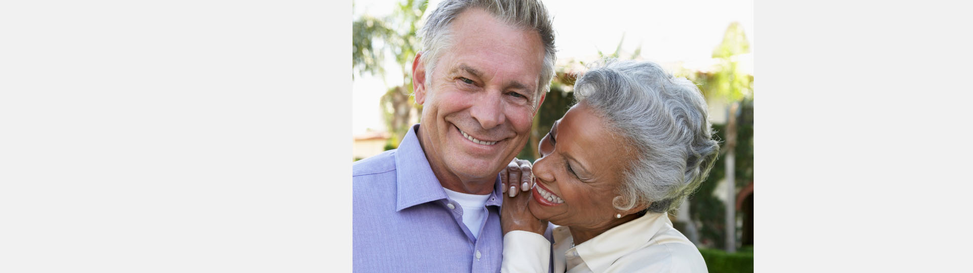 Older couple smiling and embracing in a sunny outdoor garden.