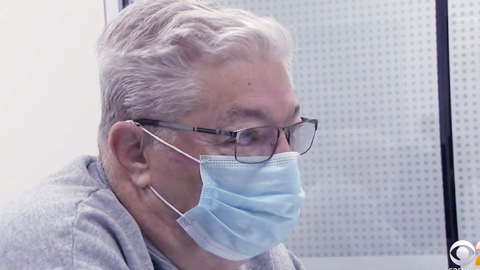 An older adult wearing glasses and a medical face mask, seated indoors near a perforated wall, looking attentively to the left.