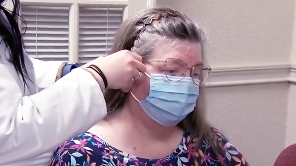 A person wearing a floral-patterned blouse is fitted with a surgical mask by a healthcare professional in a room with white walls and shuttered windows.