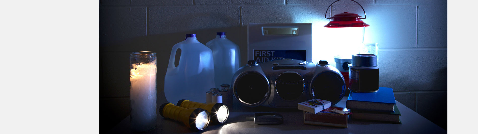 Emergency preparedness supplies for power outages, featuring lanterns, flashlights, radio, candles, water jugs, batteries, first aid kit, matches, and books on a table.