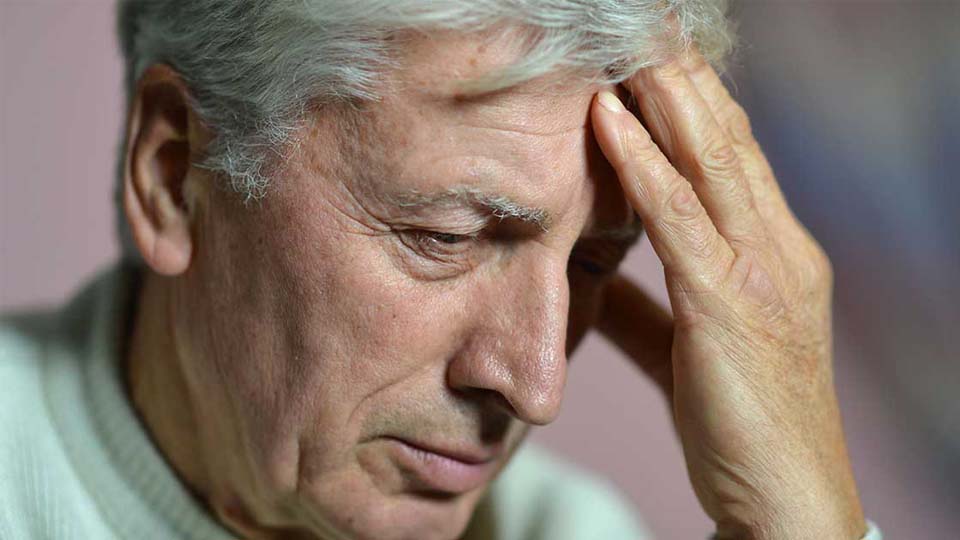 An elderly man with white hair holds his forehead with his hand, appearing thoughtful or concerned, in a softly lit indoor environment with a blurred background.