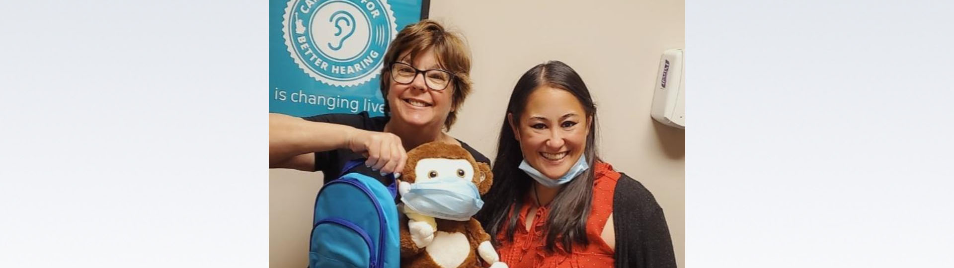 Vicki and a colleague in a hearing care clinic holding a stuffed animal with a face mask.