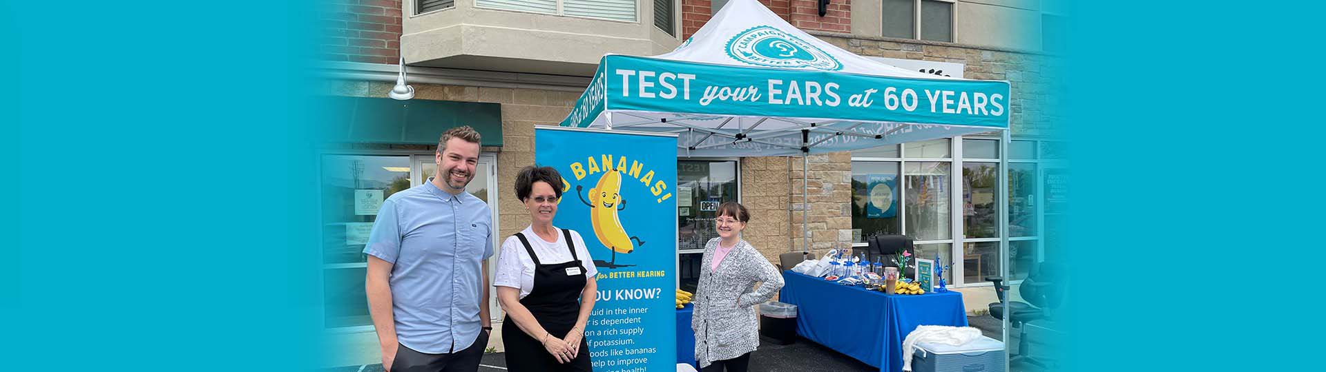 Three people standing outside under a tent with signage promoting hearing tests for individuals aged 60 and above.