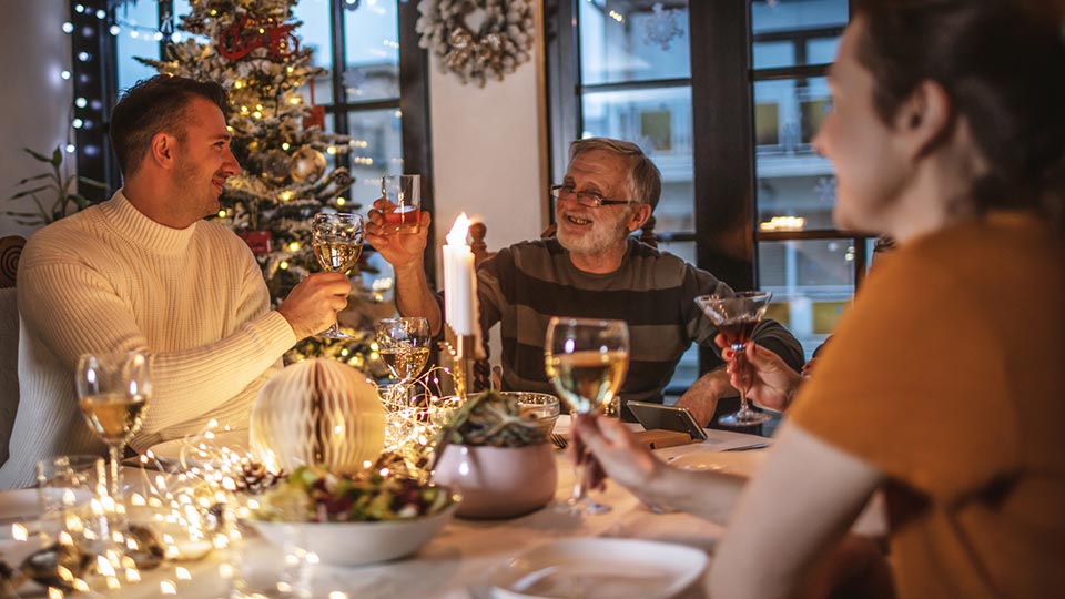 Family celebrating the holidays with food, drinks, and conversation around a festive dining table decorated with warm lights and a Christmas tree in the background.