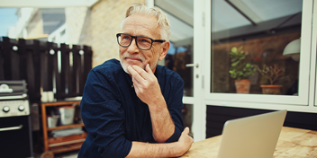 Older man sitting outdoors, thoughtful, with a laptop on a wooden table near a glass door and potted plants.