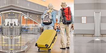 Older couple traveling through an airport with backpacks and a yellow suitcase.