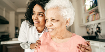 Older woman with white hair in a pink shirt being assisted by a caretaker in a cozy home environment.