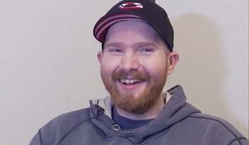 A smiling man wearing a black baseball cap and gray hoodie sits against a neutral beige wall.
