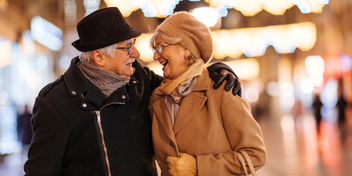Older couple smiling warmly at each other outdoors at night, wearing winter coats and hats with glowing festive lights in the background.