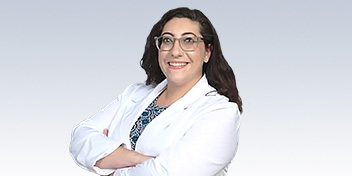 Dr. Eleni Santarelli, Audiologist at HearingLife, wearing a white coat with arms crossed, standing against a light gradient background