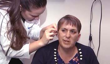 A healthcare professional adjusts a woman's hearing aid in a clinical setting with beige walls and a black wire visible in the background.