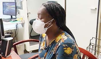 A person wearing a mask seated at a desk in an office, with a computer, chair, and filing basket visible in the background.