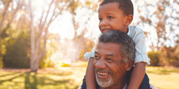 Man smiling while carrying a child on his shoulders in a sunny park.