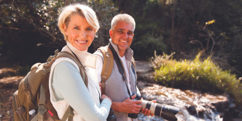 couple hiking near a stream, both wearing backpacks and hearing aids, smiling and enjoying nature