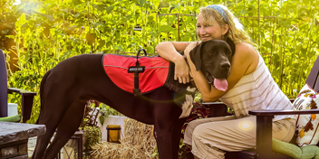 Woman embracing a hearing assistance dog wearing a red vest in a bright, sunlit garden.