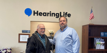 HearingLife community event with Jerry and Hearing Instrument Specialist Mike Massie standing in front of the HearingLife logo in a professional setting.