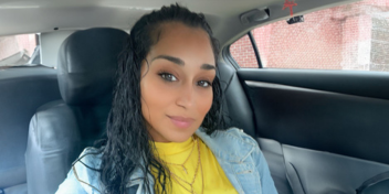 Smiling woman seated inside a car wearing a yellow shirt and denim jacket.