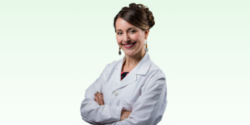 Professional woman in a white lab coat on a light green gradient background, smiling confidently with arms crossed.