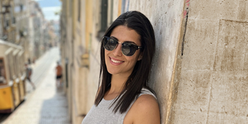 woman with dark hair wearing sunglasses, smiling on a sunny day with a tram and cobblestone urban street in the background