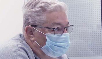 A person with gray hair and glasses, wearing a blue surgical mask, is seated indoors near a perforated white panel, possibly in a clinic or workspace.