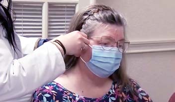 A woman in a floral blouse and glasses sits while a healthcare worker adjusts her blue surgical mask in a clinical setting with louvered windows.