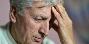 An elderly man touches his forehead with his right hand, looking pensive or worried, with a softly blurred indoor background.