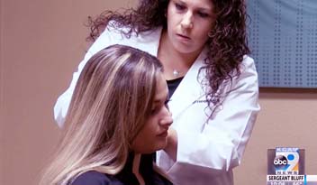 A healthcare professional examines a seated patient's scalp in a clinic with beige walls. Visible on-screen text includes a news station logo.