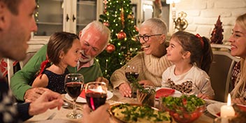 Family gathered for dinner, with grandparents and grandchildren sharing smiles and conversations around a table set with wine, festive food, and warm Christmas decorations, including a lit tree in the background.