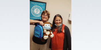 Vicki holding a stuffed monkey with a mask and a blue backpack, standing beside a colleague in a hearing care center.