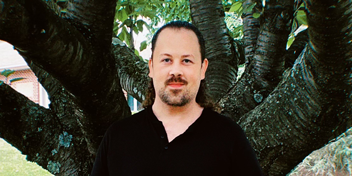 Standing outdoors in front of a large tree trunk with lush green leaves.