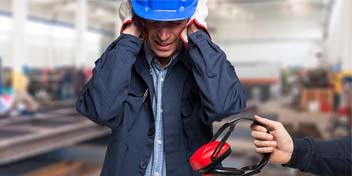 A worker in a blue hard hat covers their ears with their hands in a factory setting as ear protection is offered to them.