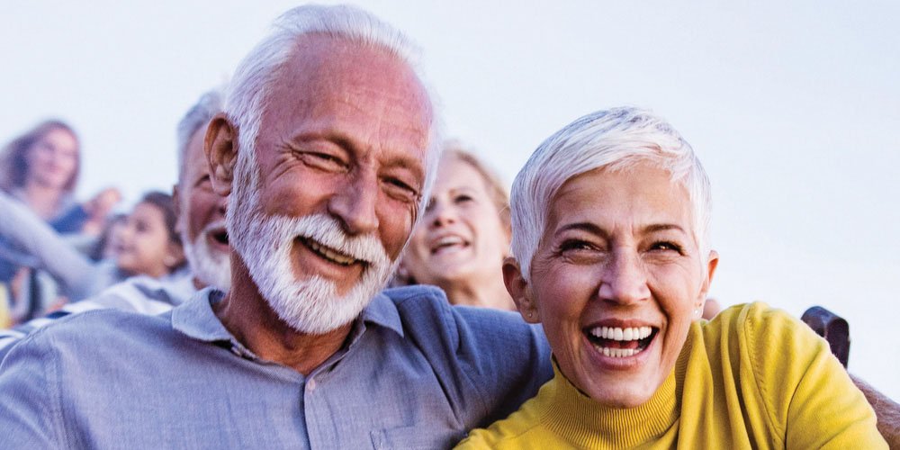 An elderly couple with grey and white hair are laughing together in an outdoor setting, surrounded by smiling people in the background.
