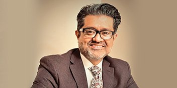 Mario Mendez wearing a brown suit jacket and patterned tie against a neutral background.