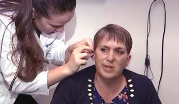A healthcare professional in a white coat adjusts a woman's hearing aid in a clinical setting, with beige walls and a black wire hanging in the background.