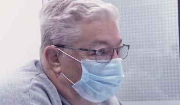 A person with gray hair and glasses, wearing a blue surgical mask, sits indoors, possibly at a work desk or clinic, with a perforated white panel behind them.